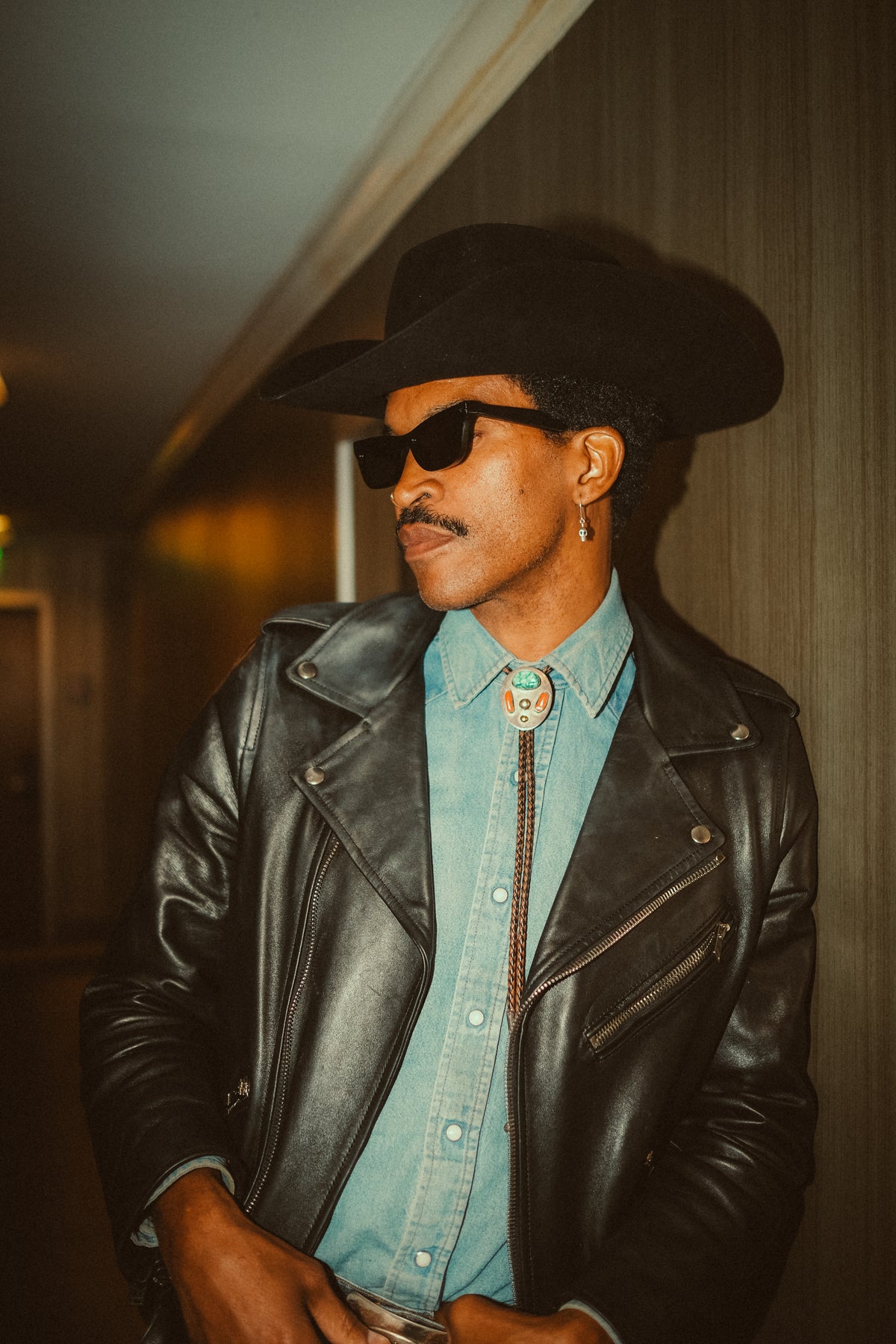 Model wearing black sunglasses, a black hat and leather jacket.